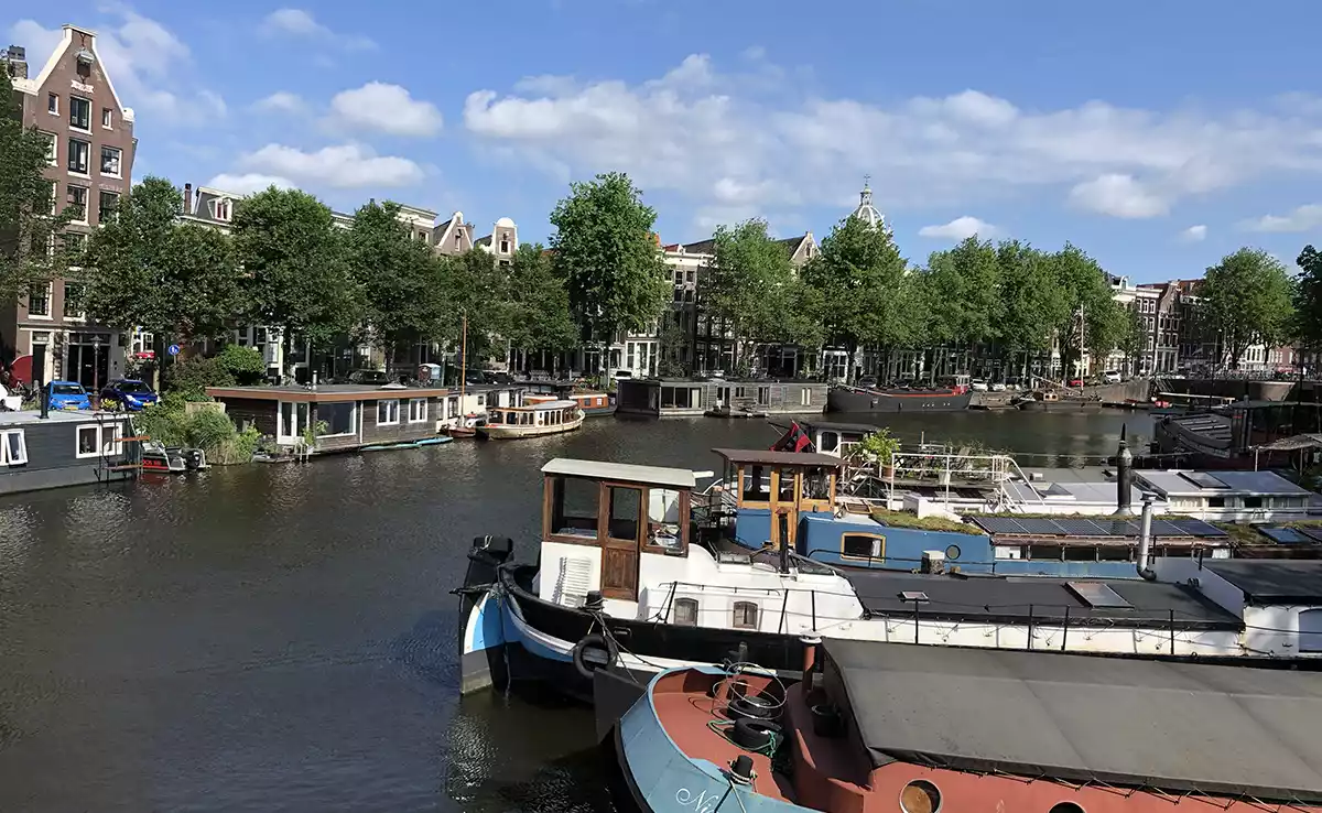 View of a canal on a sunny day, with boats in the foreground, and buildings and trees stretching along the far bank